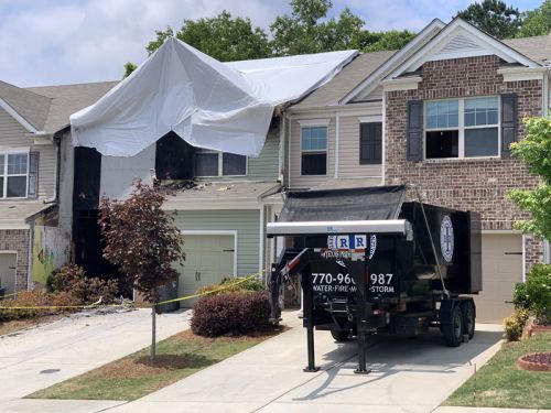 Fire-damaged townhouse covered with protective tarps during restoration work, with a debris trailer parked in the driveway and neighboring homes unaffected.